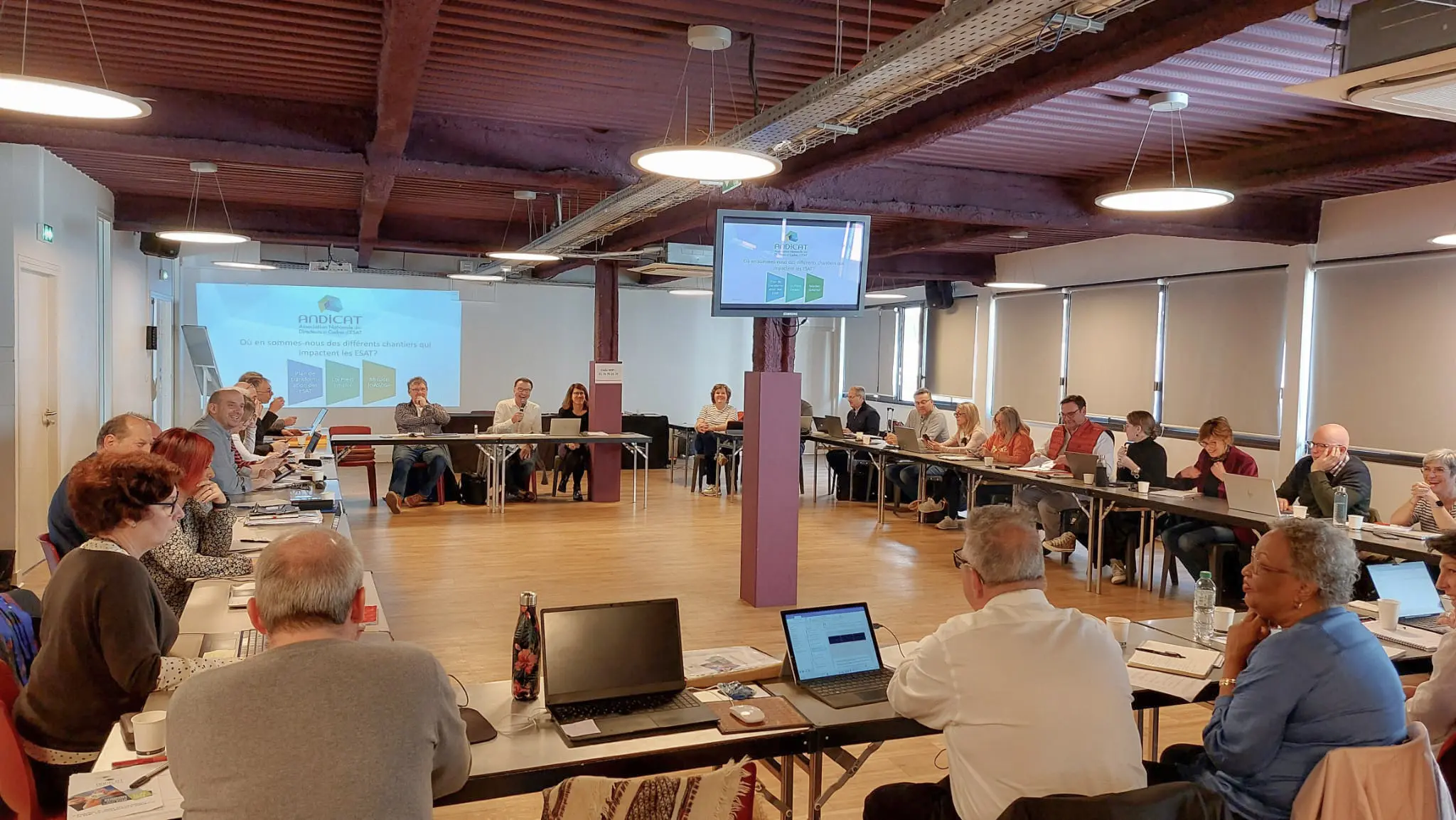 Réunion plénière d’un réseau professionnel, participants assis en cercle autour de tables avec présentation projetée à l’écran.